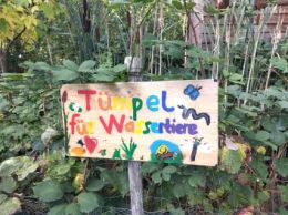Bemaltes Holzschild mit der Aufschrift „Tümpel für Wassertiere“ vor einem naturnah bepflanzten Gartenbereich.
